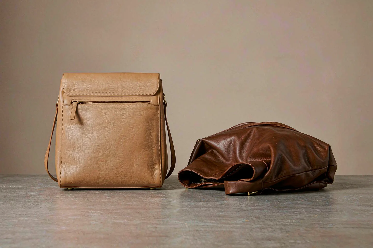 A structured tan leather satchel standing upright next to a slouchy brown leather hobo that has collapsed onto its side
